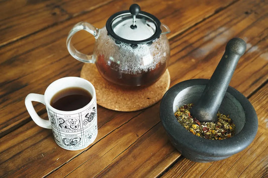 A MORTAR AND PESTLE FILLED WITH DRIED CHAMOMILE AND ROSEHIPS. A RICH BROWN WOODEN COUNTERTOP IS UNDER AND THE MORTAR AND PESTLE SITS NEXT TO A POT OF FRESHLY BREWED TEA IN A CLEAR GLASS TEAPOT. TO THE LEFT OF BOTH IS A MUG FILLED WITH A EARTHY BROWN TEA IN A 1970S WHITE ASTROLOGY MUG WITH A SMALL CHIP IN THE BOTTOM CENTER. 